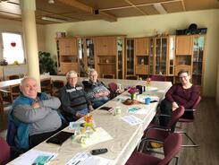 An einem l&auml;ngeren Tisch sitzen auf der linken Seite zwei Senioren und eine Seniorin, auf der rechten Seite sitzt eine Frau. Die vier Personen gucken nach vorne in die Kamera. Auf dem Tisch liegen verschiedenen Informationsmaterialien.