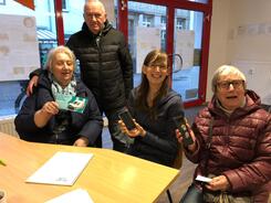 Mobilreferentin Petra (Mitte) sitzt mit einer Seniorin (rechts) und einem &auml;lteren Ehepaar (links) an einem Tisch. Petra und die rechte Seniorin halten ein Smartphone in die Kamera. Die Ehefrau h&auml;lt den Flyer des Digitalen Engel.