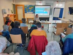 Mobilreferentin Theresa sitzt an einem Tisch und zeigt den Teilnehmenden Doctolib. Im Hintergrund ist die Doctolib-Internetseite auf einer Leinwand zu sehen. Die Teilnehmerinnen sitzen auf St&uuml;hlen und sind von hinten zu sehen. 