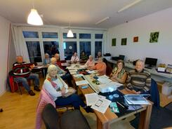 In einem Raum im MGH Buntes Haus sitzen vier &auml;ltere M&auml;nner und acht &auml;ltere Frauen an einem l&auml;nglichen Tisch. Die meisten blicken nach vorne in die Kamera. Auf dem Tisch im vorderen Bildrand stehen ein Beamer und ein Laptop.