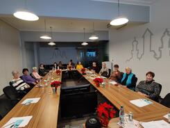 14 Seniorinnen und Senioren sitzen in einem gro&szlig;en Seminarraum im Stadtarchiv an einem langen Tisch in U-Form. Sie gucken nach vorne in die Kamera. Auf dem Tisch liegen Winterdekorationen und Unterlagen vom Digitalen Engel.