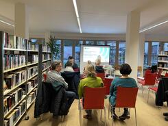 Die Teilnehmenden sitzen in einer Bibliothek auf roten St&uuml;hlen und sind von hinten zu sehen. Sie schauen auf die Titelfolie zum Thema hilfreiche Apps im Alltag &auml;lterer Menschen.