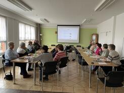 Die Teilnehmenden sitzen an zwei l&auml;ngeren Gruppentischen und schauen auf die Leinwand, auf der die Stationen des Digitalen Engels angezeigt werden. Die Wand hinter der Leinwand ist hellgr&uuml;n.