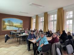 Die Teilnehmenden sitzen sich gegen&uuml;ber an drei Gruppentisch. Einige sind von hinten zu sehen, andere l&auml;cheln in die Kamera.