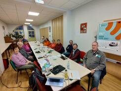 An einem langen Tisch sitzen dreizehn Senior:innen und blicken nach vorne in die Kamera. Am Bildrand rechts h&auml;ngt ein Plakat vom Digitalen Engel mit Informationen zu der Veranstaltung.