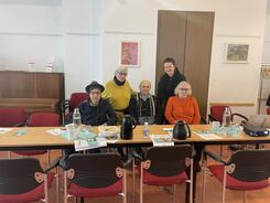 3 Teilnehmende sitzen auf schwarz-roten St&uuml;hlen, zwei Teilnehmerinnen stehen hinter ihnen. 