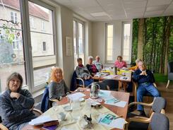 An einem Tisch im Vordergrund sitzen zwei Seniorinnen, an einem hinteren Tisch sitzen vier Frauen und zwei M&auml;nner. Auf den Tischen liegen Materialien wie der Flyer vom Digitalen Engel.
