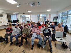 &Uuml;ber zwanzig j&uuml;ngere und &auml;ltere Senior:innen sitzen in vier Stuhlreihen im DIZ und blicken nach vorne in die Kamera.
