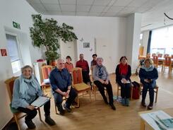 Acht Senior:innen (zwei M&auml;nner und sechs Frauen) sitzen in einem Raum auf St&uuml;hlen und l&auml;cheln nach vorne in die Kamera.