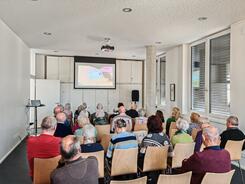 Mehrere Personen sitzen auf St&uuml;hlen im Raum und blicken nach Vorne auf eine Leinwand auf der ein Video abgespielt wird.