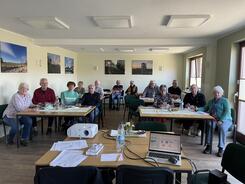 Die 13 Teilnehmenden sitzen an Tischen mit bis zu vier Personen und l&auml;cheln in die Kamera.