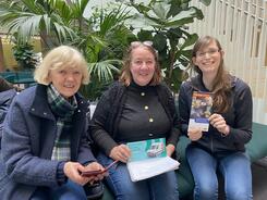 Petra (rechts) sitzt mit den beiden Freundinnen Astrid (Mitte) und Christine (links) im Atrium des K&ouml;rberHauses. Petra und Astrid halten zwei Flyer in den H&auml;nden und Christine ihr Smartphone.