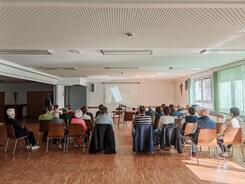 Zahlreiche Personen sitzen auf St&uuml;hlen im Raum verteilt und blicken nach Vorne zu einer Leinwand.