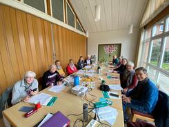 An einem gro&szlig;en, langen Tisch sitzen elf Senior:innen, davon ein Mann. Auf dem Tisch im vorderen Bildrand steht ein Beamer.