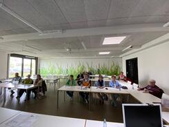In einem gro&szlig;en Raum sitzen insgesamt 15 Menschen. Das Foto ist im Weitwinkel-Modus aufgenommen, damit alle Teilnehmende, die im Raum verteilt sitzen, abgebildet sind.