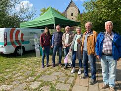 Petra (links) steht mit f&uuml;nf Senior:innen (vier M&auml;nner und eine Frau) vor dem Info-Mobil und einem gr&uuml;nen Pavillon.