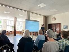 Mehrere Personen sitzen auf St&uuml;hlen im Raum und blicken nach Vorne zu einer Leinwand und zu Referent Johannes.