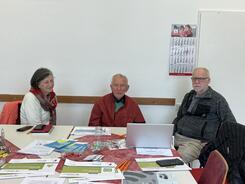 Die drei Teilnehmenden sitzen an einem mit Brosch&uuml;ren und Handouts gedeckten Tisch und l&auml;cheln in die Kamera.