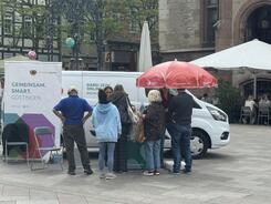 Aus der Ferne ist der Info-Stand mit dem Info-Mobil fotografiert. Es stehen mehrere Passant:innen an dem Stand und sind im Gespr&auml;ch. 