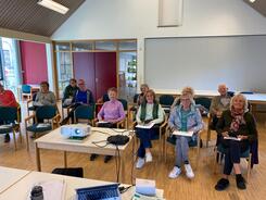 Im Dorftreff Ankum sitzen in einem Raum auf St&uuml;hlen elf Personen und blicken in die Kamera. Vor ihnen steht ein Tisch mit einem Beamer.