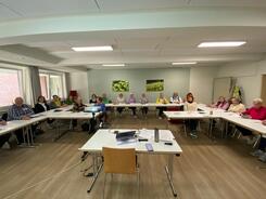 Ein Schulungsraum mit einer Gruppe &auml;lterer Menschen, die in einem U-f&ouml;rmig angeordneten Sitzkreis an Tischen sitzen. Alle Teilnehmenden schauen freundlich in die Kamera. Auf den Tischen liegen Unterlagen, Stifte und digitale Ger&auml;te. Im Vordergrund ist ein Laptop und ein Beamer zu sehen.