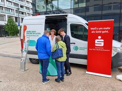 Johannes steht neben einem Berater der Sparkasse vor dem Infomobil und unterh&auml;lt sich mit einem Ehepaar.