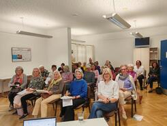 Mehrere Personen sitzen neben und hintereinander auf St&uuml;hlen im Raum und blicken nach vorne.