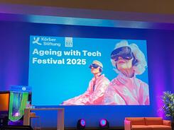 Eine B&uuml;hne im Theatersaal des K&ouml;rberHauses. Eine Pr&auml;sentation zeigt eine Folie mit der &Uuml;berschrift "Ageing with Tech Festival 2025". Rechts ist eine KI-generiertes Bild mit einer &auml;lteren Dama und einer j&uuml;ngeren Person, die eine VR-Brille tragen.