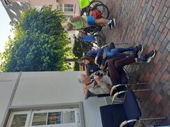 Auf dem Bild im Hochkantformat sitzt Petra mit einem &auml;lteren Herr auf zwei St&uuml;hlen. Sie erkl&auml;rt ihm etwas auf dem Smartphone. Rechts im Hintergrund steht ein sommerlich gekleideter Mann an einem Fahrrad.