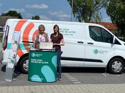 Petra (rechts) steht mit Seniorin Johanne (links) am Info-Stand vor dem Info-Mobil. Petra h&auml;lt den Laptop von Johanne.