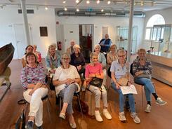 In einem Museumraum sitzen knapp f&uuml;nfzehn Senior:innen in drei Stuhlreihen und schauen nach vorne in die Kamera.
