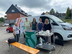 Am Info-Mobil steht Petra (Mitte) mit Anna (2. v. r.) und Maren Korban von der AWO (1. v. r.). Links von Petra steht ein AWO-Vorstandsmitglied und Annas Stammg&auml;stin. Vor den f&uuml;nf Personen steht der Info-Stand mit einer Bierbank und einem Wagen, auf dem Kaffee und Wasser steht.