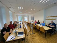 In einem l&auml;nglichen Raum sitzen an zwei langen Tisch knapp 12 Personen. Auf den Tischen liegen Materialien und Getr&auml;nke. Zwischen den Tischen steht ein Beamer auf einer Erh&ouml;hung.