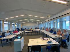 In einem sehr gro&szlig;en Raum sitzen ca. 13 Personen an Tischen, die in einer U-Form aufgestellt sind. Im Vordergrund steht ein Tisch mit einem Laptop und ein kleiner Schrank mit einem Beamer.