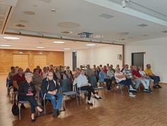 Zahlreiche Personen sitzen auf St&uuml;hlen in einem gro&szlig;en Saal und blicken nach Vorne.