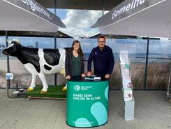 Petra (links) steht mit B&uuml;rgermeister J&uuml;rgen Schiewer (rechts) am Info-Stand vor einem Supermarkt. Links neben Petra steht eine lebensgro&szlig;e Kuh-Figur.