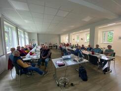 Das Foto ist im Weitwinkelmodus aufgenommen. Es zeigt &uuml;ber drei&szlig;ig Personen, die an Tischen in U-Form sitzen. Im Vordergrund steht ein Tisch mit einem Laptop und Beamer.