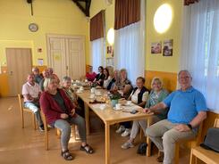 In der DRK-Sozialstation sitzen zehn Senior:innen und die drei Sch&uuml;lerinnen an einem langen Tisch und gucken nach vorne in die Kamera. Auf dem Tisch liegen Materialien und Kaffeegedeck.