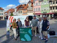 Petra (dritte von links) steht mit  f&uuml;nf Senioren, darunter Mitglieder des Seniorenbeirats, an dem Info-Stand auf dem Marktplatz in Helmstedt. Im Hintergrund sind historische H&auml;user zu sehen.
