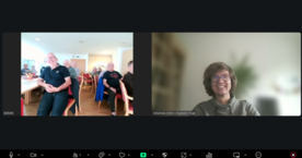 Bildschirmfoto vom Zoom-Fenster, in dem links die Teilnehmenden im Raum sitzend zu sehen sind und rechts Referent Johannes l&auml;chelnd in die Kamera blickt.