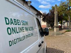 Links im Vordergrund ist das Info-Mobil mit folgender Aufschrift abgebildet: "Dabei sein - Online im Alter! digitaler-engel.org". Rechts im Hintergrund steht die Posts&auml;ule.