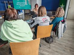 Drei Frauen sitzen auf St&uuml;hlen an einem Tisch und blicken nach Vorne auf einen Bildschirm und zum Referenten, der am Laptop sitzt.