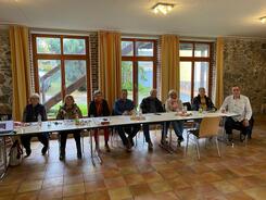 An einem langen Tisch sitzen acht Personen (drei M&auml;nner und f&uuml;nf Frauen) und gucken nach vorne in die Kamera.