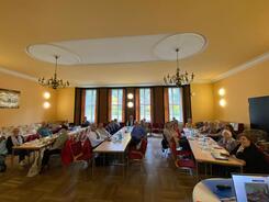 In einem gro&szlig;en Saal sitzen an drei langen Tischen insgesamt knapp 30 Senior:innen.