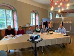 In einem Raum sitzen an einem Tisch sechs Senior:innen (vier Frauen in der Mitte und zwei M&auml;nner an den R&auml;ndern). In der Mitte steht ein Beamer.