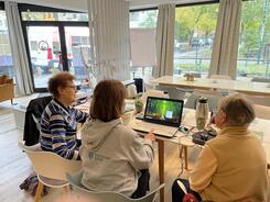 Theresa und zwei Teilnehmerinnen sitzen an einem Tisch und sind von hinten zu sehen. Theresa zeigt den beiden &auml;lteren Frauen Dinge am Laptop.