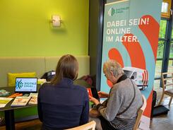 Petra (links) sitzt mit einer &auml;lteren Dame (rechts) an einem Tisch. Die beiden sind von hinten fotografiert und blicken aus ein Smartphone. Rechts von den beiden ist ein Roll Up mit der Aufschrift "Dabei sein! Online im Alter". Link steht ein Tablet mit dem Engel-Logo.