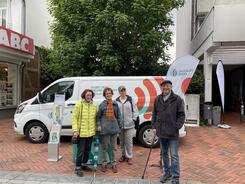 4 Besucher:innen stehen in einer Einkaufsstra&szlig;e vor dem Infomobil am Infostand und l&auml;cheln in die Kamera. Der Himmel ist dunkel aufgrund bevorstehenden Regens. 