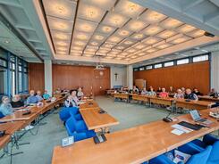 Zahlreiche Personen sitzen an mehreren zusammengeschobenen Tischen und blicken nach Vorne.
