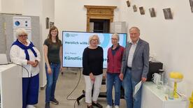 Petra (zweite von links) steht mit Barbara Schmidt vom Seniorenbeirat (erste von links) mit drei weiteren Teilnehmenden vor einem Bildschirm, auf dem "Herzlich willkommen" steht mit dem Logo des Digitalen Engel.